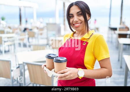 Junge schöne arabische Kellnerin lächelt selbstbewusst und hält Kaffee zum Mitnehmen auf der Terrasse des Cafés Stockfoto