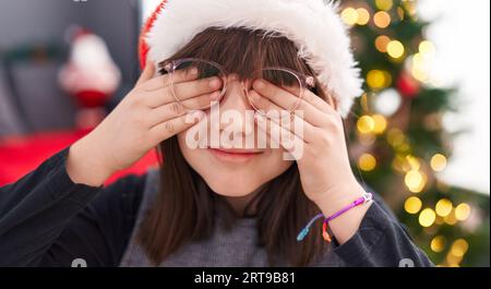 Entzückendes hispanisches Mädchen, das die Augen mit Händen bedeckt, die zu Hause am weihnachtsbaum stehen Stockfoto