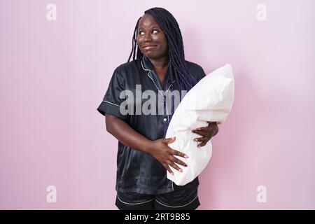 Eine junge afrikanerin, die ein Pijama trägt und ein Kissen umarmt, lächelt, schaut zur Seite und schaut nachdenklich weg. Stockfoto