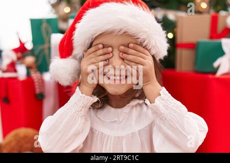 Entzückendes hispanisches Mädchen, das die Augen mit Händen bedeckt, die zu Hause am weihnachtsbaum auf dem Boden sitzen Stockfoto