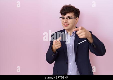 Junger, nicht binärer Mann mit Bart, der Anzug trägt und mit den Fingern auf die Kamera zeigt, mit fröhlichem und lustigem Gesicht. Gute Energie und gute Stimmung. Stockfoto