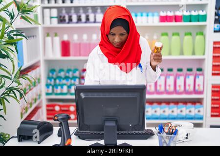 Eine junge, schöne Apothekerin, die einen Computer benutzt und eine Pillenflasche in der Apotheke hält Stockfoto