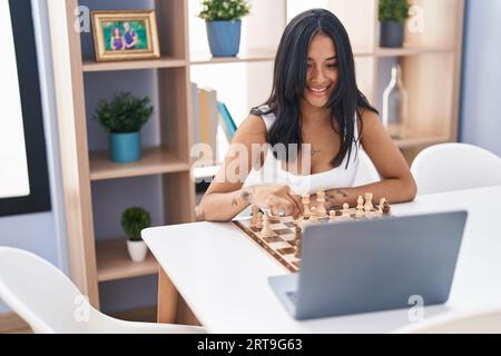 Junge hispanische Frau, die online Schach spielt, sitzt zu Hause auf einem Tisch Stockfoto