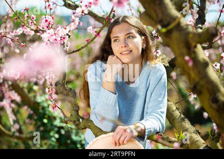 Lächelndes Mädchen in blauem Pullover, das unter dem Blütenbaum steht Stockfoto