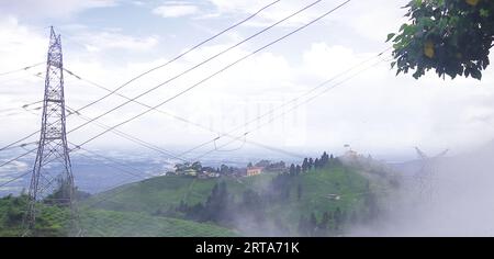 Hochspannungs-Stromverteilungsnetzmasten, die in der Regenzeit durch die Berge vor den Ausläufern und das Tal mit bewölktem bedecktem Hintergrund führen Stockfoto