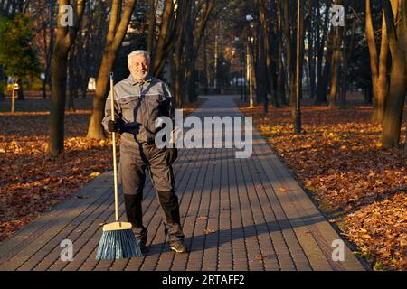 Reifer Arbeiter, der vor der Kamera posiert, mit Besen im Park. Vorderansicht eines glücklichen bärtigen Mannes im Alter, in Uniform stehend mit Besen und lächelnd, mit Stockfoto