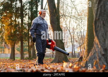 Älterer Mann, der im Herbst mit Laubbläser die Straße aufräumt. Schwacher Blick auf den fokussierten bärtigen Mann, der im Stadtpark arbeitet und Blätter in Morni entfernt Stockfoto