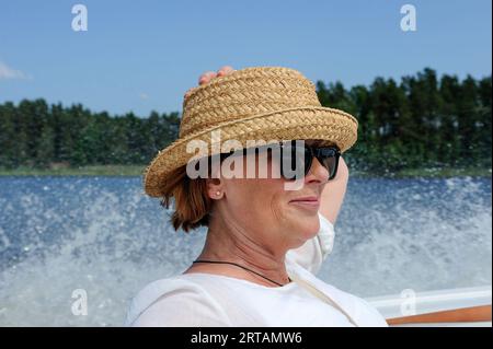 Frau im Profil, mit der Hand auf dem Strohhut, mit Sonnenbrille, die in einem Boot sitzt und dahinter Wasser spritzt Stockfoto