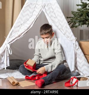 Weihnachten Mit Der Familie. Kinder öffnen Weihnachtsgeschenke. Ein Teenager öffnet ein Geschenk und lächelt, während er auf dem Boden eines Hauses sitzt, das für Neujahr dekoriert ist Stockfoto