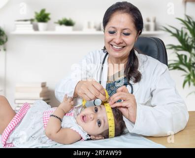 Lächelnde Kinderärztin untersucht 6 Monate kleines Mädchen und misst das Wachstum. Arzt mit Maßband zur Überprüfung der Kopfgröße des Babys. Neugeborenes Routin Stockfoto