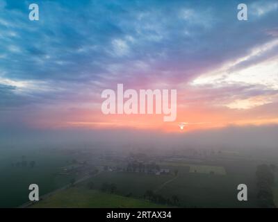 Ein beeindruckendes Luftpanorama, das die Schönheit eines nebeligen, bewölkten morgens einfängt, während die Sonne über einer ländlichen Landschaft aufgeht. Verschiedene Felder, jedes mit verschiedenen Ernten, erstrecken sich so weit das Auge reicht, gebadet in das warme, bunte Licht der Morgenröte. Majestätischer Sonnenaufgang Über Ländlicher Landschaft. Hochwertige Fotos Stockfoto