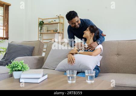 Glücklicher junger Mann umarmt Frau von hinten auf dem Sofa, nachdem sie von der Arbeit nach Hause gekommen ist Stockfoto