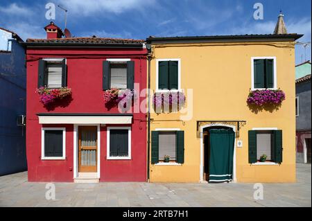 Hellrote und gelbe Häuser mit frischen Blumen an den Fenstern, die sich am bewölkten blauen Himmel auf der Straße der Insel Burano in Venedig, Italien, befinden Stockfoto