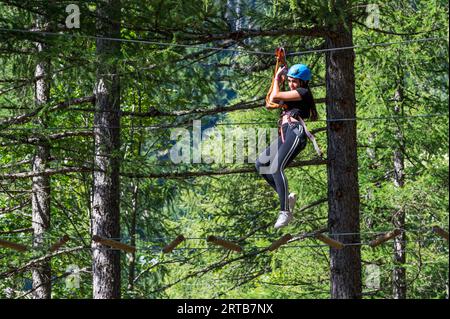 Niedriger Winkel, Seitenansicht der aufgeregten jungen Frau im Helm, während sie am Oberseil mit Sicherheitsseilen hängt und auf einem Hängedraht im Wald geht Stockfoto