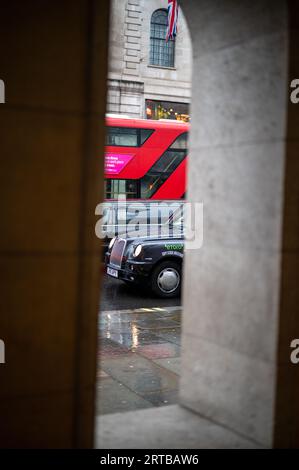 LONDON - 24. April 2023: Eine klassische Szene Londons: Ein roter Doppeldeckerbus und ein schwarzes Taxi, eingerahmt von einem Bogen an einem regnerischen Tag, der das britische U verkörpert Stockfoto