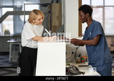 Seitenansicht des jungen afroamerikanischen männlichen Klinikers, der weiblichen Klienten hilft, das medizinische Dokument zu unterzeichnen, während er sich über den Administratorzähler beugt Stockfoto