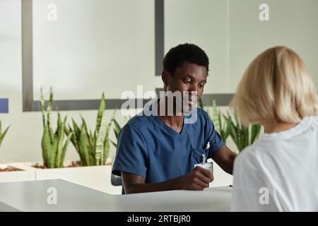 Junger afroamerikanischer Kliniker, der eine Patientin anschaut, die ein medizinisches Dokument unterzeichnet, während sie vor ihm an der Rezeption steht Stockfoto