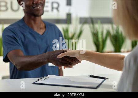 Fokus auf Handschlag von jungen Arzt oder Verwalter in blauer Uniform und Patientin über Empfangstheke mit unterzeichnetem medizinischen Dokument Stockfoto