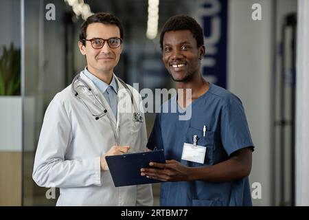 Zwei glückliche junge interkulturelle Kliniker in Uniform stehen vor der Kamera und schauen Sie an, während sie über das medizinische Dokument diskutieren Stockfoto