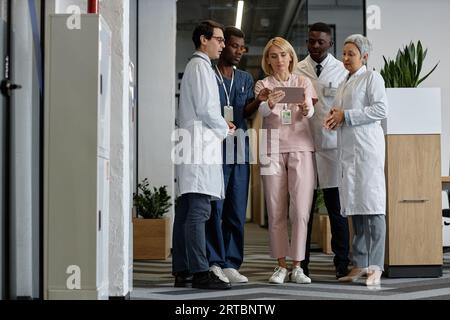 Fünf interkulturelle Ärzte und Assistenten in Uniform, die medizinische Daten auf dem Tablet-Bildschirm diskutieren, während sie im geräumigen Korridor des Krankenhauses stehen Stockfoto