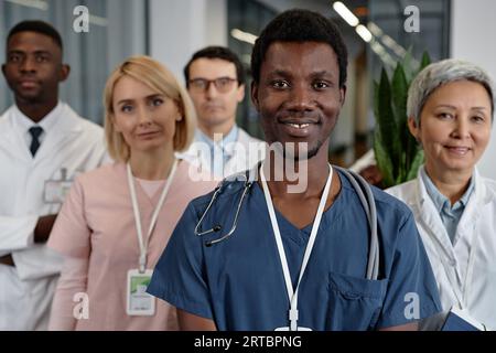 Junge lächelnde afroamerikanische männliche Assistentin in blauer Uniform mit Blick auf die Kamera, während sie gegen eine Gruppe interkultureller Kollegen steht Stockfoto