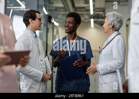 Glücklicher junger afroamerikanischer männlicher Assistent mit medizinischem Dokument, das mit Kollegen spricht und ihnen Arbeitspunkte beim Treffen erklärt Stockfoto