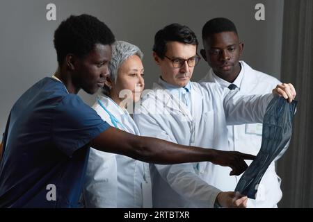 Junger, selbstbewusster afroamerikanischer Mann in blauer Uniform, der auf das Röntgenbild des Patienten zeigt, während er es mit einer Gruppe von Kollegen beim Treffen diskutiert Stockfoto