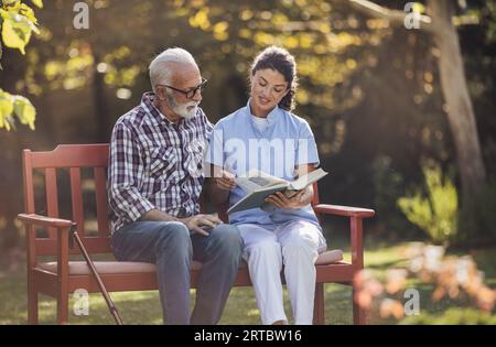 Junge Pflegekräfte, die auf einer Bank im Park sitzt und dem älteren Mann mit Gehstock Buch liest Stockfoto