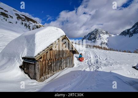 Frau auf einer Skitour sitzt auf einer schneebedeckten Alm mit Bielschitza im Hintergrund, Matschacher Alm, Rosental, Karawanken, Kärnten, Österreich Stockfoto