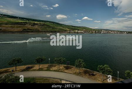 Blick über die Rheinpromenade von Bingen zu einem Hotelschiff auf dem Rhein, im Hintergrund der Niederwald und die Altstadt Rüdesheim, obere mittlere R Stockfoto