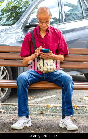 Älterer Mann in magentafarbenem Hemd auf der Bank im Stadtzentrum, Smartphone lernend - Tours, Indre-et-Loire (37), Frankreich. Stockfoto