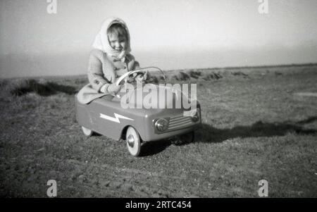 1950er Jahre, historisch, draußen auf freiem Gelände, ein kleines Mädchen mit Mantel und Kopftuch, das in einem Spielzeugwagen fährt/spielt, England, Großbritannien. Stockfoto