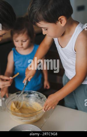 Kleiner Junge, der seiner Mutter beim Backen in der Küche hilft, die neben seiner kleinen Schwester steht und den Teig für Themuffins knetet. Konzept Liebe und Bindung. Stockfoto
