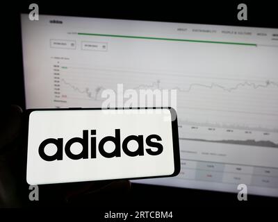 Person, die ein Mobiltelefon mit dem Logo des deutschen Bekleidungs- und Schuhherstellers Adidas AG auf dem Bildschirm vor der Webseite hält. Konzentrieren Sie sich auf das Display des Telefons. Stockfoto