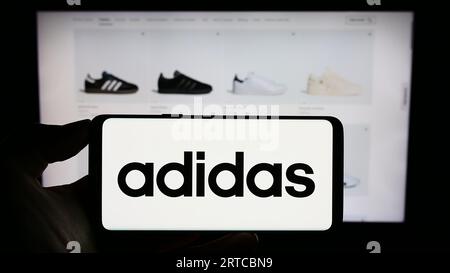 Person, die ein Mobiltelefon mit dem Logo des deutschen Bekleidungs- und Schuhherstellers Adidas AG auf dem Bildschirm vor der Webseite hält. Konzentrieren Sie sich auf das Display des Telefons. Stockfoto