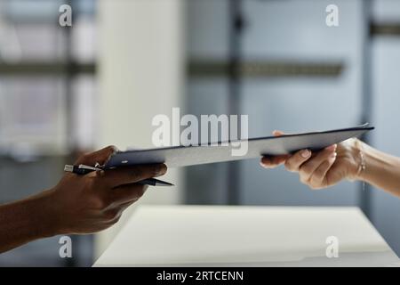 Seitenansicht der Hand einer jungen weiblichen Patientin, die das Clipboard mit dem medizinischen Dokument an einen afroamerikanischen männlichen Rezeptionisten oder Kliniker übergibt Stockfoto