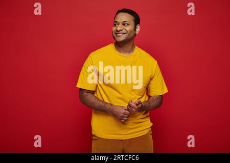 Entzückender indischer Mann in heller, lässiger Kleidung, der im Studio auf rotem Hintergrund lächelt und wegschaut Stockfoto