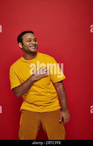 Dankbarer indischer Mann in heller Freizeitkleidung lächelnd und auf rotem Hintergrund im Studio wegschauend Stockfoto