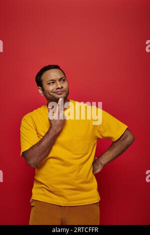 Nachdenklicher indischer Mann in heller Freizeitkleidung, der auf rotem Hintergrund wegschaut, kreative Idee Stockfoto