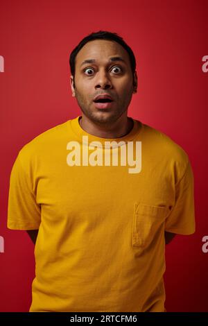 staunender indischer Mann in gelbem T-Shirt, der die Kamera auf rotem Hintergrund betrachtet, Gesichtsausdruck Stockfoto