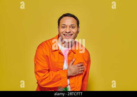 Porträt eines freudigen indischen Mannes in oranger Jacke, der auf die Kamera schaut und auf gelbem Hintergrund lächelt Stockfoto