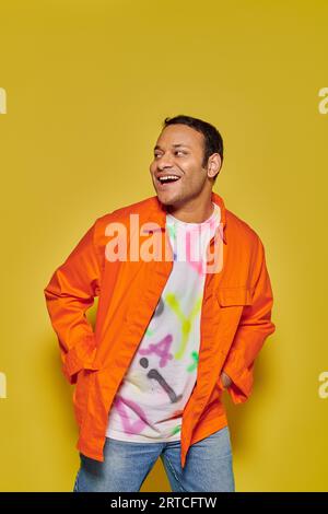 Porträt eines fröhlichen indischen Mannes in oranger Jacke mit Händen in Taschen auf gelbem Hintergrund Stockfoto