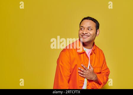 Porträt eines positiven indischen Mannes in oranger Jacke, der wegschaut und lächelt auf gelbem Hintergrund Stockfoto