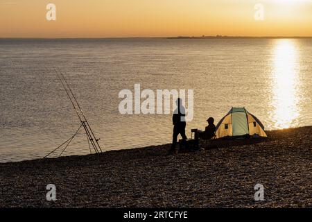 Hurst Beach, Hampshire, Großbritannien - September 2023: An einem Kiesstrand bei Sonnenuntergang, vor dem Zelt, warten zwei Angler mit ihren Ruten auf einen Happen. Stockfoto