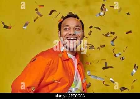 Fröhlicher indischer Mann in leuchtend oranger Jacke lächelnd nahe fallendem Konfetti auf gelbem Hintergrund Stockfoto