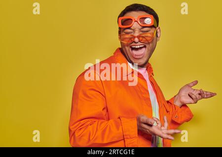 Lustiges Gesicht, aufgeregter indischer Mann, der verschiedene trendige Sonnenbrillen anprobiert und auf gelbem Hintergrund lächelt Stockfoto