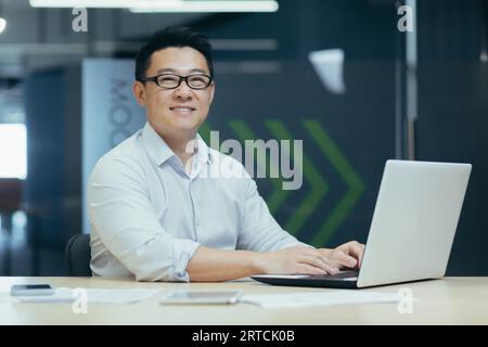 Porträt eines jungen asiatischen männlichen Architekten, Brilleningenieur, der im Büro arbeitet, am Tisch am Laptop sitzt und vor der Kamera lächelt. Stockfoto