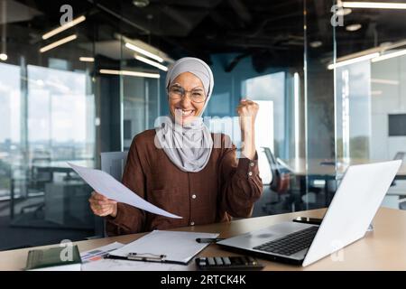 Die junge muslimische Geschäftsfrau in Hijab ist glücklich mit Erfolg, Gewinn und Abschluss. Im Büro mit Dokumenten am Tisch sitzend und mit der Hand zur Kamera eine Siegesgeste zeigend. Stockfoto