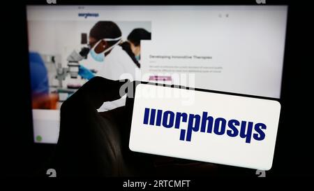 Person, die ein Mobiltelefon mit dem Logo des deutschen biopharmazeutischen Unternehmens MorphoSys AG auf dem Bildschirm vor der Webseite hält. Konzentrieren Sie sich auf das Display des Telefons. Stockfoto