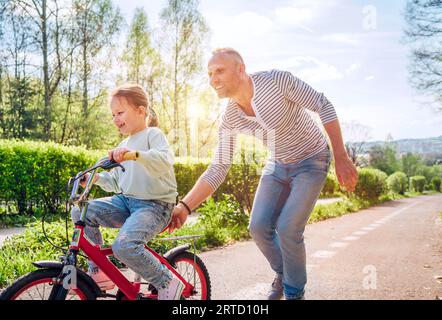 Lächelnder Vater brachte seiner Tochter Fahrradfahren bei. Sie genießen die Zusammengehörigkeit im Sommer-Stadtpark. Fröhliche Kindheit und Outdoor-Aktivitäten Stockfoto